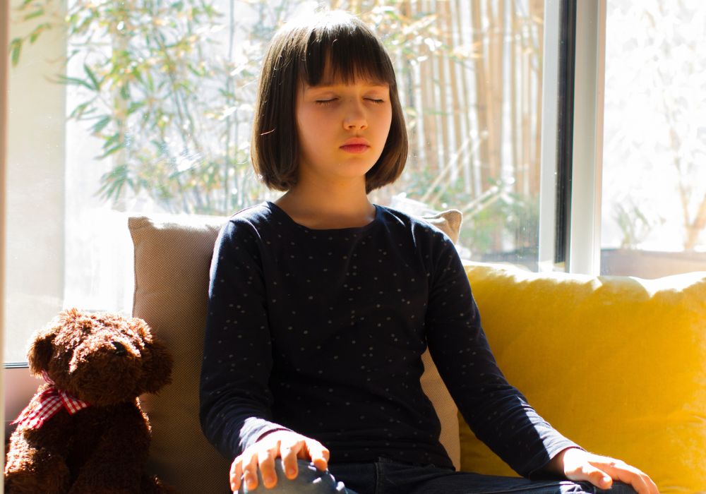 girl sitting next to teddy bear with eyes closed, practicing meditation
