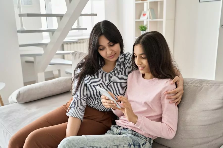 Girl being supported by mother, sitting on lounge looking at phone
